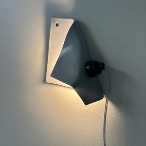 May include: A wall-mounted light fixture featuring a curved, light gray metal shade that directs illumination downward. The fixture includes a black socket, a visible power cord, and a small mounting hole at the top of the flat metal backplate.