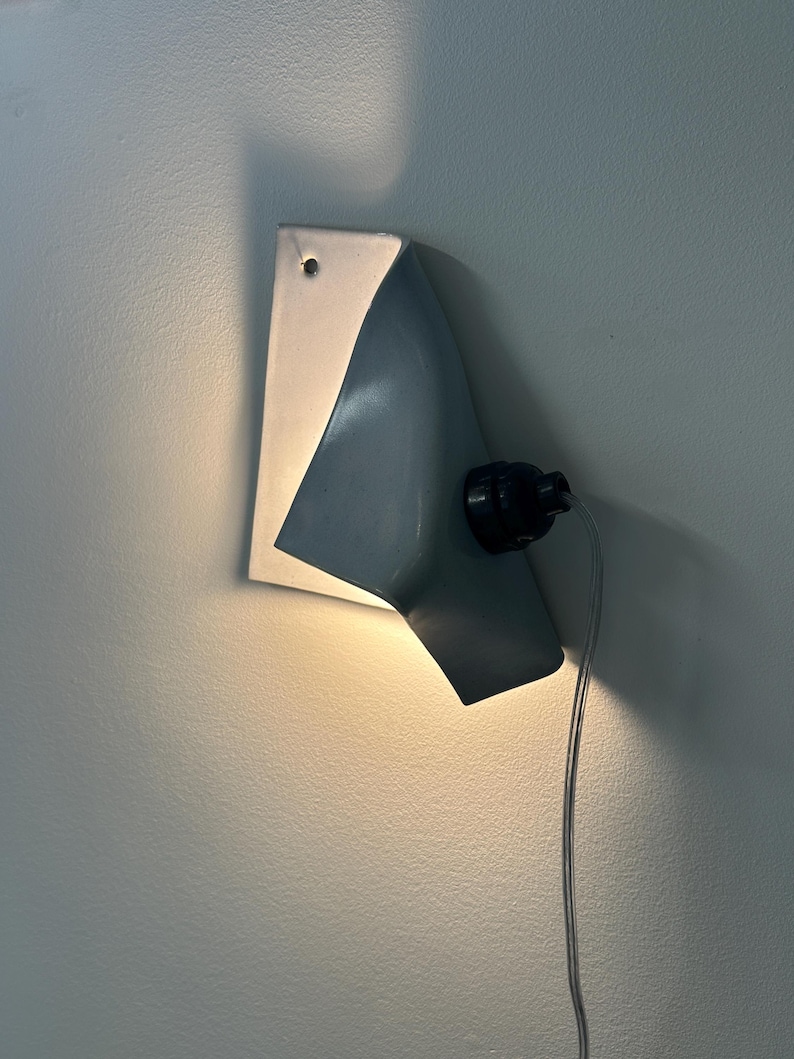 May include: A wall-mounted light fixture featuring a curved, light gray metal shade that directs illumination downward. The fixture includes a black socket, a visible power cord, and a small mounting hole at the top of the flat metal backplate.