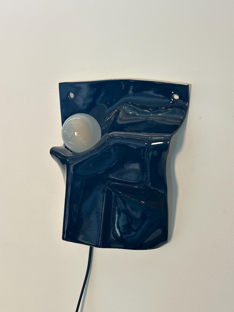Puede incluir: Un aplique de pared de cer&aacute;mica azul oscuro con una superficie brillante y ondulada y una bombilla blanca redonda. El accesorio cuenta con dos peque&ntilde;os orificios de montaje circulares en la parte superior y un cable negro en la base.