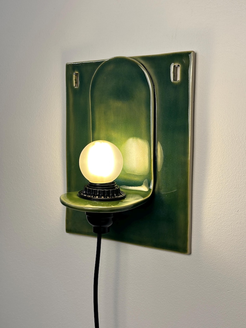 May include: A wall-mounted ceramic sconce featuring a glossy dark green finish with a rectangular backplate and a curved shelf. A small, frosted spherical light bulb sits on the shelf, connected to a black cord extending downward from the base.