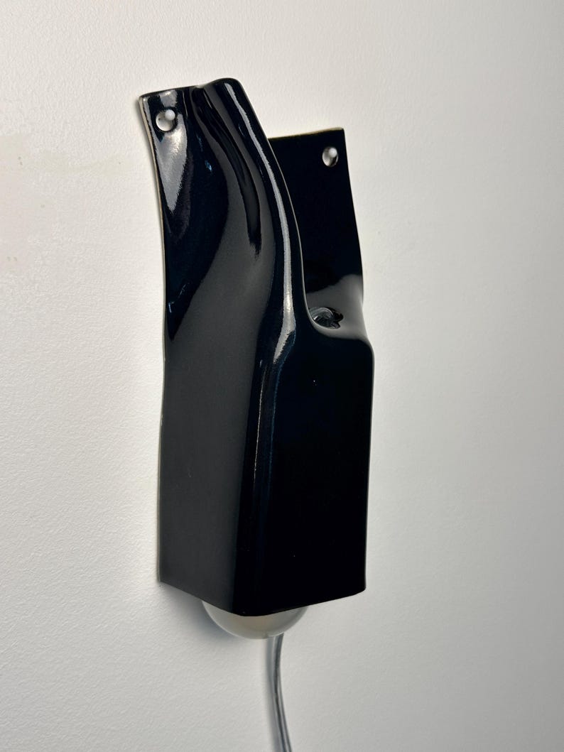 May include: A wall-mounted lamp featuring a glossy, dark navy blue ceramic fixture with a curved, organic silhouette. The structure includes two visible mounting holes at the top and a small, round, white light bulb protruding from the bottom.