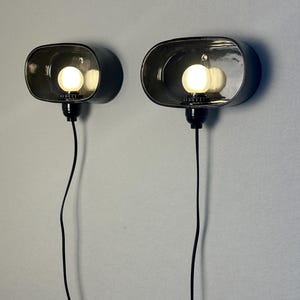 May include: A pair of wall-mounted lamps with an oval, dark grey shade. Each lamp has a bright, round bulb. Black cords hang from the base. The lamps are fixed to a light grey wall. The lamps are approximately 20 cm in length.