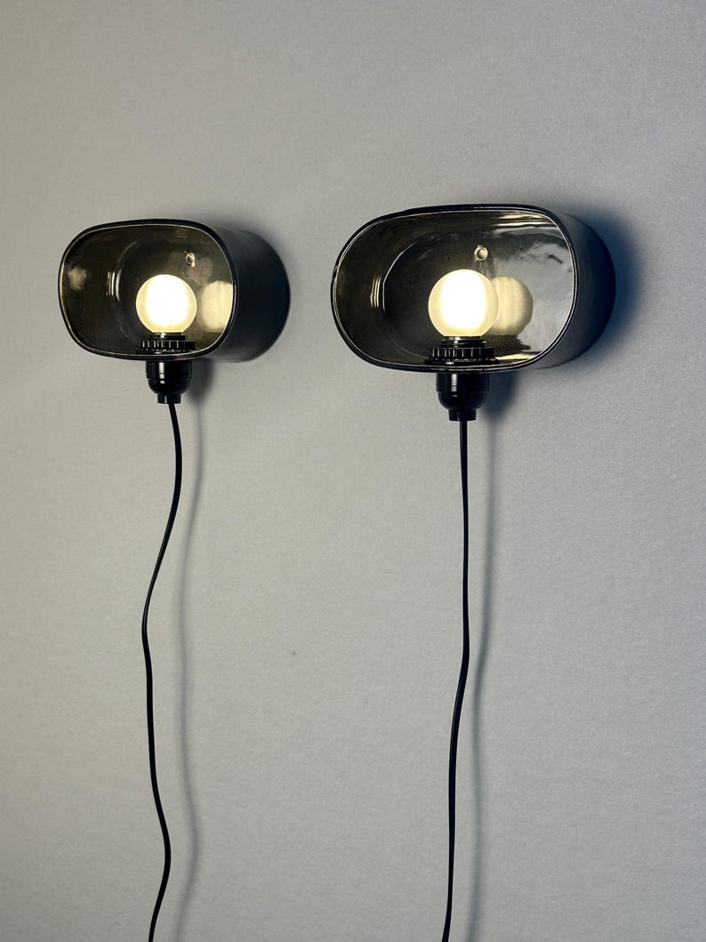 May include: A pair of wall-mounted lamps with an oval, dark grey shade. Each lamp has a bright, round bulb. Black cords hang from the base. The lamps are fixed to a light grey wall. The lamps are approximately 20 cm in length.