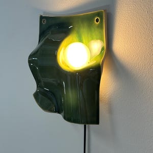May include: A wall-mounted lamp featuring a glossy, undulating ceramic shade in shades of deep forest green and bright lime. The organic, wave-like form surrounds a central, glowing spherical light bulb, with a black power cord extending downward.