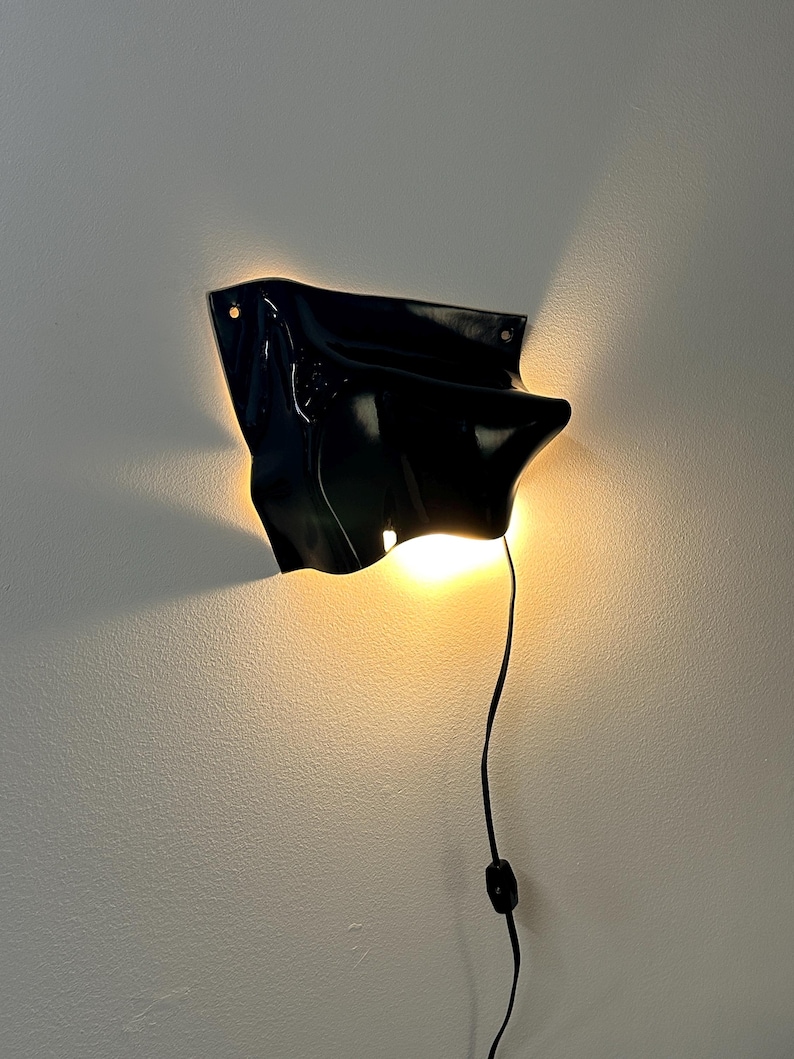 Puede incluir: Un aplique de pared negro con una superficie irregular, plegada y brillante. La l&aacute;mpara emite una luz c&aacute;lida y resplandeciente desde la parte inferior, proyectando haces suaves contra la pared. Un cable negro con interruptor cuelga abajo.