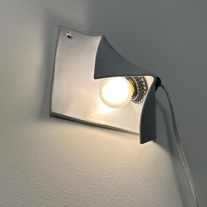 May include: A white wall-mounted lamp with a curved, folded sheet design. The fixture features a single glowing light bulb positioned within the concave fold, and a thin, transparent electrical cord extending from the side of the light fixture.