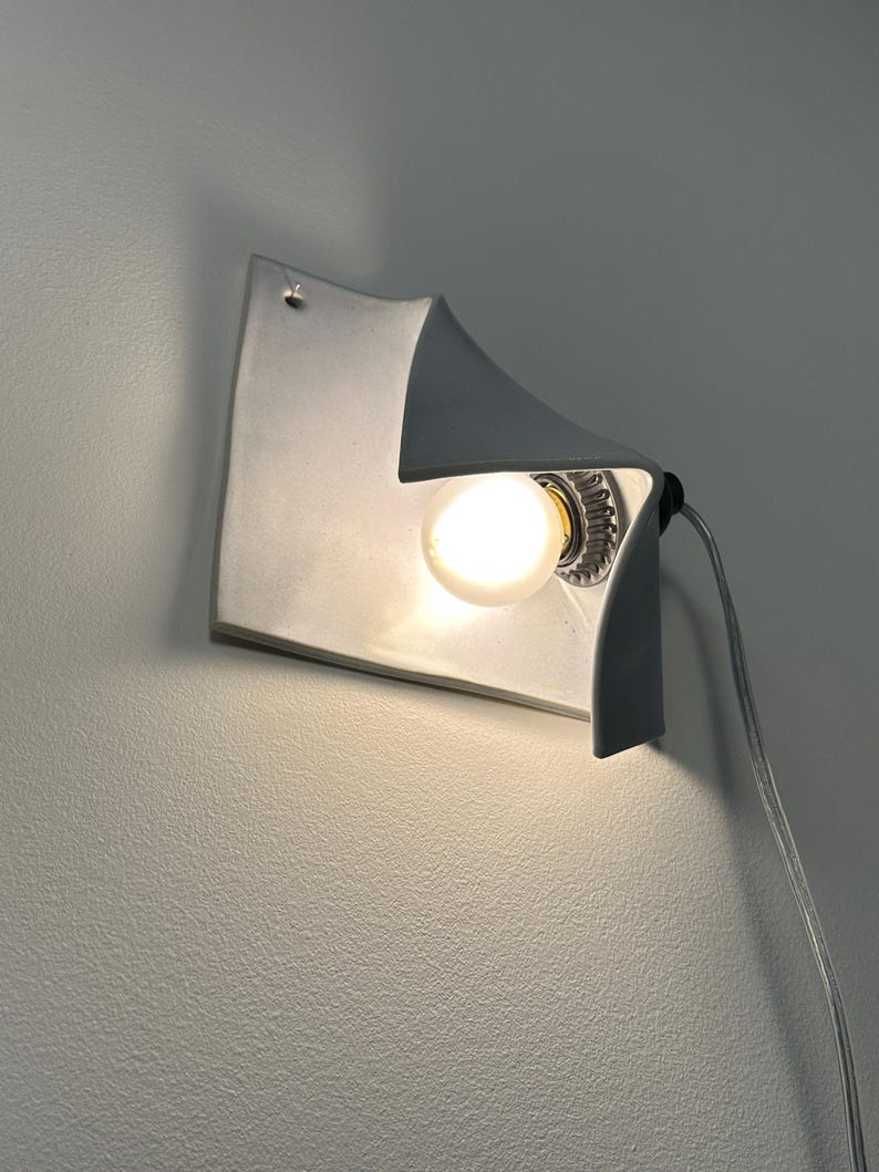 May include: A white wall-mounted lamp with a curved, folded sheet design. The fixture features a single glowing light bulb positioned within the concave fold, and a thin, transparent electrical cord extending from the side of the light fixture.