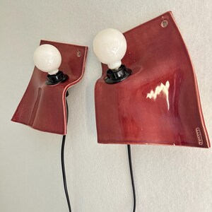 Puede incluir: Dos l&aacute;mparas de pared con placas traseras de cer&aacute;mica roja brillante, curvas y de forma ondulada y org&aacute;nica. Cada l&aacute;mpara tiene un casquillo negro con una bombilla blanca redonda y un cable de alimentaci&oacute;n negro. El texto TALLERDAD es visible.