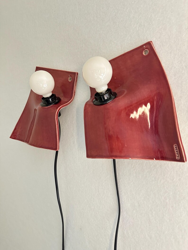 Puede incluir: Dos l&aacute;mparas de pared con placas traseras de cer&aacute;mica roja brillante, curvas y de forma ondulada y org&aacute;nica. Cada l&aacute;mpara tiene un casquillo negro con una bombilla blanca redonda y un cable de alimentaci&oacute;n negro. El texto TALLERDAD es visible.