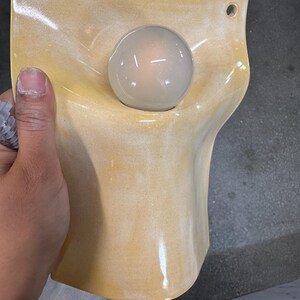 May include: A wall-mounted light fixture featuring a curved, pale yellow ceramic shade with a glossy finish. The shade has two circular holes at the top for mounting and a central opening that holds a single, round, frosted white light bulb.