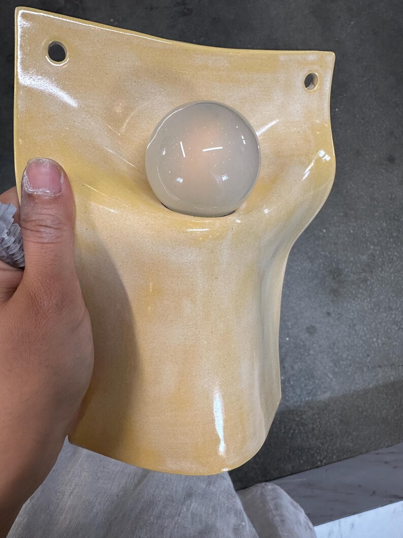 May include: A wall-mounted light fixture featuring a curved, pale yellow ceramic shade with a glossy finish. The shade has two circular holes at the top for mounting and a central opening that holds a single, round, frosted white light bulb.