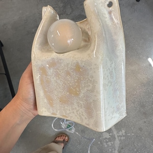 May include: A cream-coloured ceramic wall lamp with a glossy, mottled glaze and a sculptural, bag-like shape. The fixture features a circular opening at the top for mounting and a round, glowing light bulb nestled within the upper section.