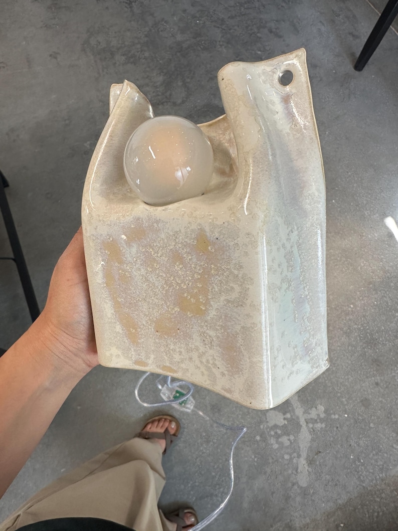 May include: A cream-coloured ceramic wall lamp with a glossy, mottled glaze and a sculptural, bag-like shape. The fixture features a circular opening at the top for mounting and a round, glowing light bulb nestled within the upper section.