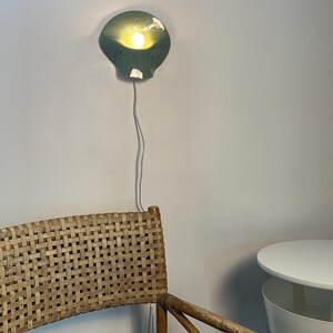 May include: A sage green wall-mounted lamp with a curved, shell-like shade emitting a warm, glowing light. The lamp features a thin white power cord extending downward. The fixture is mounted on a plain wall above a woven rattan chair.