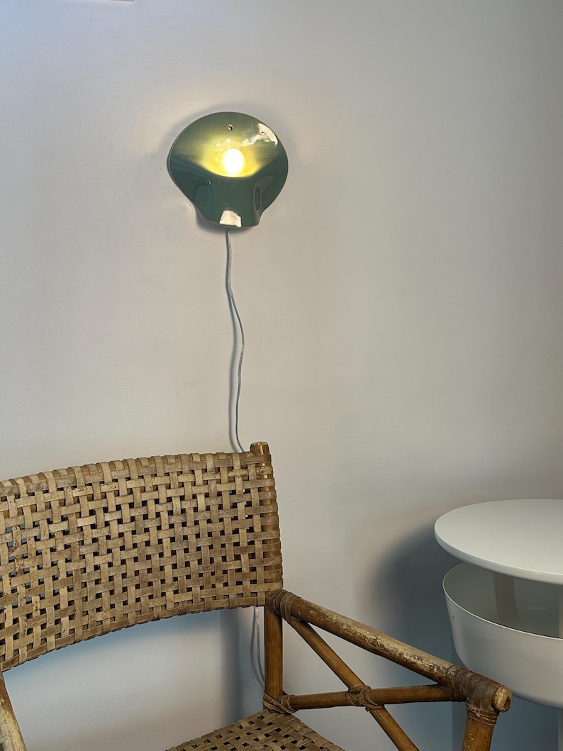 May include: A sage green wall-mounted lamp with a curved, shell-like shade emitting a warm, glowing light. The lamp features a thin white power cord extending downward. The fixture is mounted on a plain wall above a woven rattan chair.