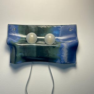 May include: A wall-mounted light fixture featuring a rectangular, wavy ceramic panel with a blue and dark green glazed finish. Two spherical, frosted white glass bulbs are mounted horizontally on the front, with two thin power cords extending downward.