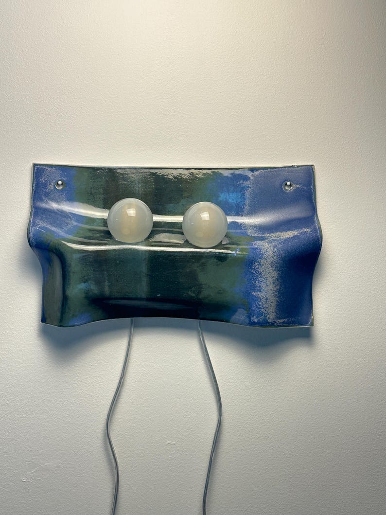 May include: A wall-mounted light fixture featuring a rectangular, wavy ceramic panel with a blue and dark green glazed finish. Two spherical, frosted white glass bulbs are mounted horizontally on the front, with two thin power cords extending downward.