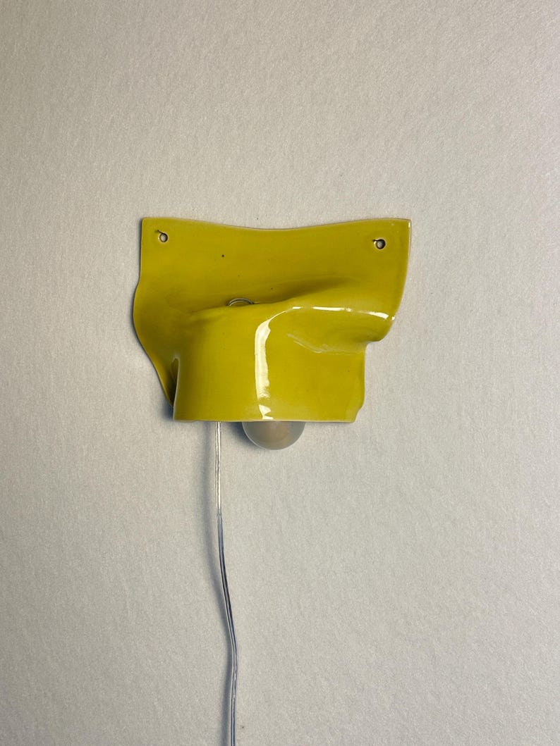 May include: A yellow ceramic wall sconce with a glossy finish. The sconce has a unique, folded design and features a small light bulb suspended by a wire. Two small holes are visible near the top for mounting.