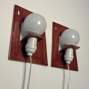Puede incluir: Un par de apliques de pared con placas traseras rectangulares de cer&aacute;mica roja oscura brillante con un centro recortado. Cada aplique tiene un casquillo blanco montado en un peque&ntilde;o estante de cer&aacute;mica, una bombilla redonda y un cable.