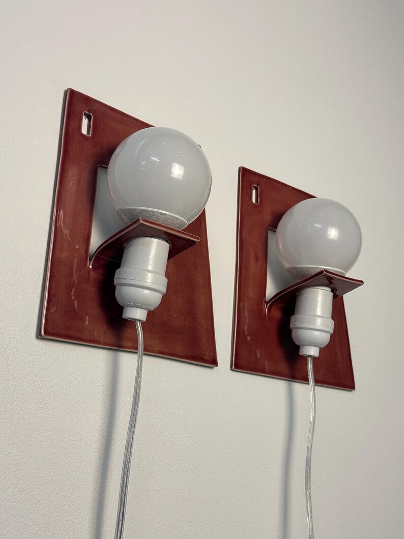 Puede incluir: Un par de apliques de pared con placas traseras rectangulares de cer&aacute;mica roja oscura brillante con un centro recortado. Cada aplique tiene un casquillo blanco montado en un peque&ntilde;o estante de cer&aacute;mica, una bombilla redonda y un cable.