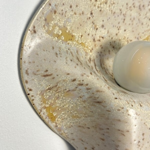 May include: A wall-mounted ceramic light fitting with a curved, organic shape. The surface features a speckled cream and beige glaze with crystalline patterns. A single, smooth, opaque white glass globe bulb is centred on the curved ceramic base.