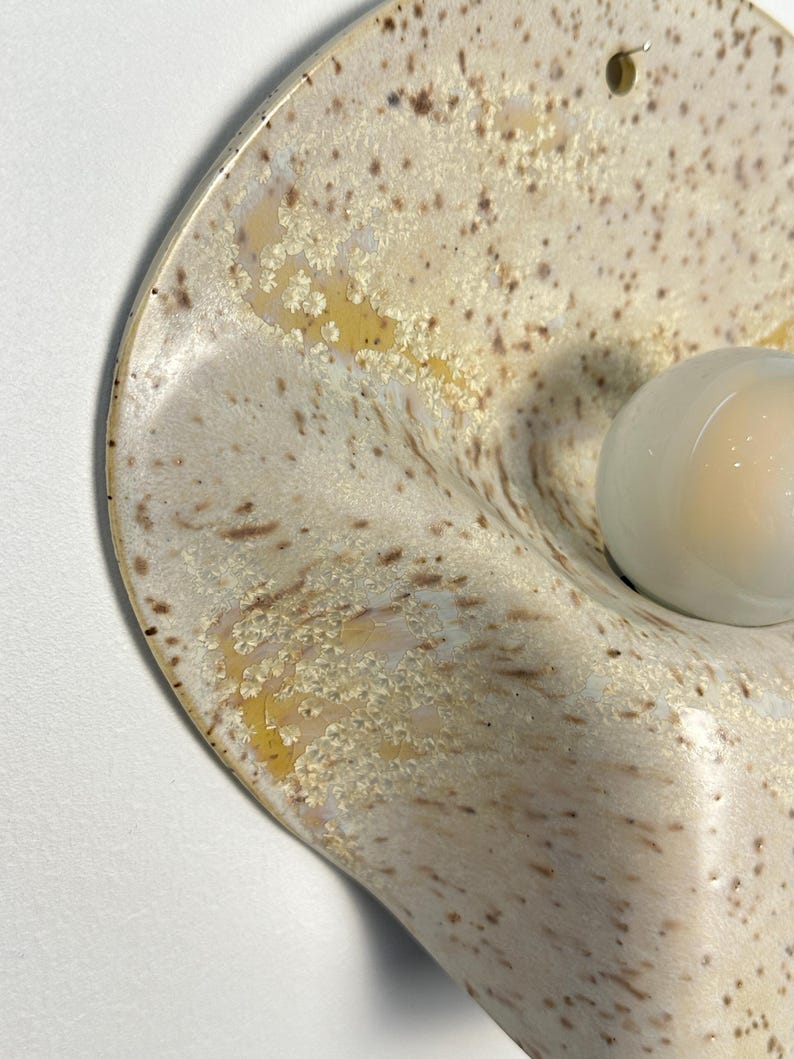May include: A wall-mounted ceramic light fitting with a curved, organic shape. The surface features a speckled cream and beige glaze with crystalline patterns. A single, smooth, opaque white glass globe bulb is centred on the curved ceramic base.