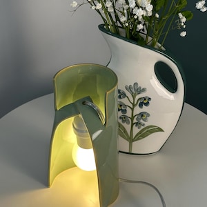 May include: A sage green ceramic table lamp with a curved, open-sided cylindrical shade and a single glowing light bulb. Beside it sits a white ceramic pitcher featuring a green rim and a painted blue floral design filled with white flowers.