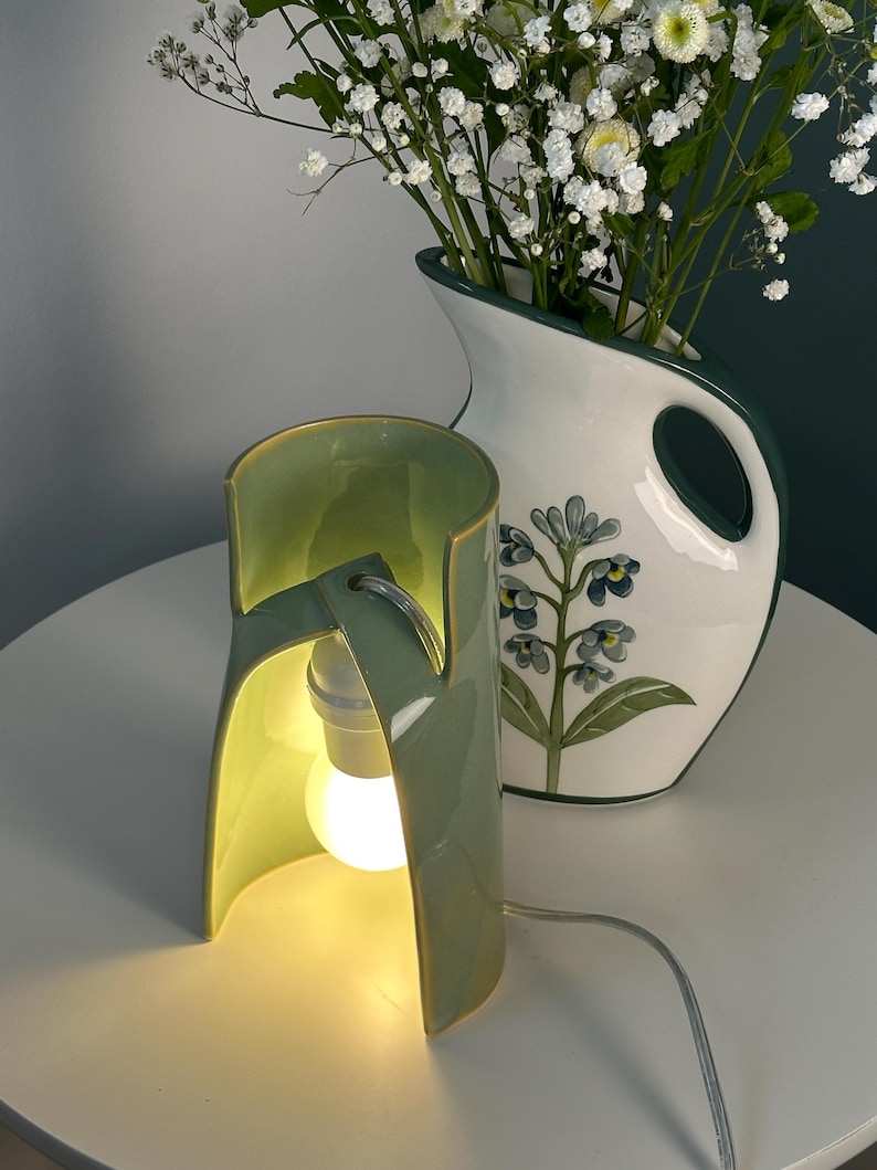 May include: A sage green ceramic table lamp with a curved, open-sided cylindrical shade and a single glowing light bulb. Beside it sits a white ceramic pitcher featuring a green rim and a painted blue floral design filled with white flowers.