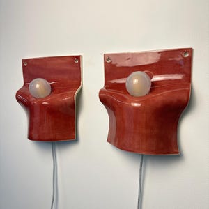 May include: Two wall-mounted sconces featuring glossy, deep red ceramic plates with organic, undulating curves. Each sconce has a central circular opening holding a frosted white globe light bulb and a grey electrical cord extending from the bottom.
