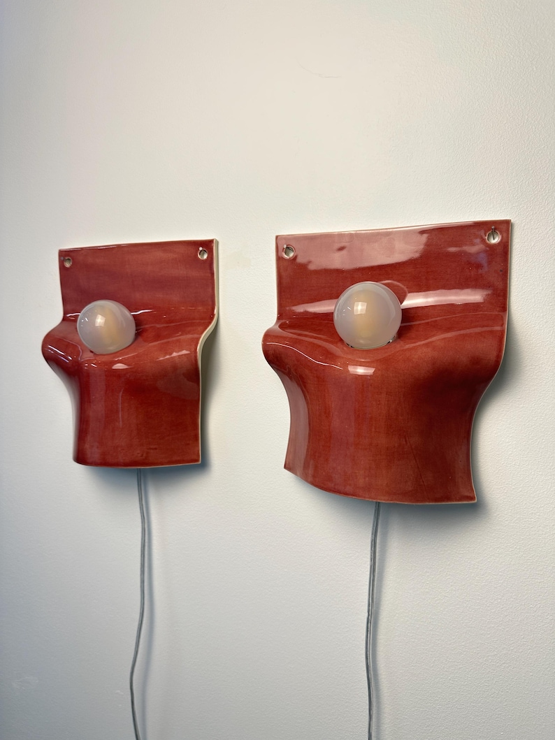 May include: Two wall-mounted sconces featuring glossy, deep red ceramic plates with organic, undulating curves. Each sconce has a central circular opening holding a frosted white globe light bulb and a grey electrical cord extending from the bottom.