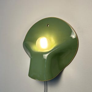 May include: A wall-mounted lamp featuring a smooth, sage green, shell-shaped ceramic shade with a glossy finish. A single, round, frosted light bulb is centered within the curved form, emitting a warm glow. A thin, light-colored power cord hangs below.