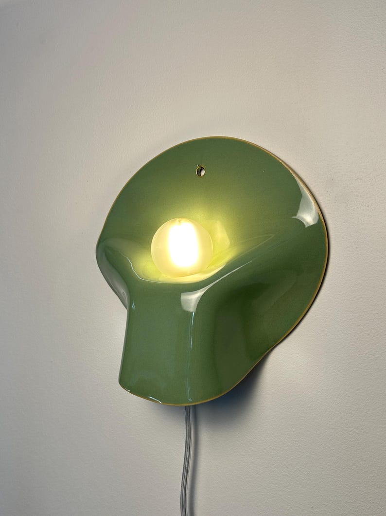 May include: A wall-mounted lamp featuring a smooth, sage green, shell-shaped ceramic shade with a glossy finish. A single, round, frosted light bulb is centered within the curved form, emitting a warm glow. A thin, light-colored power cord hangs below.