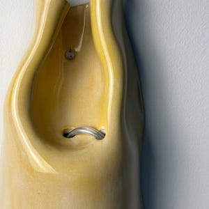 Pu&ograve; includere: Un vaso in ceramica giallo da parete con finitura lucida e forma organica ripiegata. L'oggetto presenta una piccola apertura circolare vicino alla parte superiore e un'apertura centrale pi&ugrave; ampia contenente un anello metallico argentato.