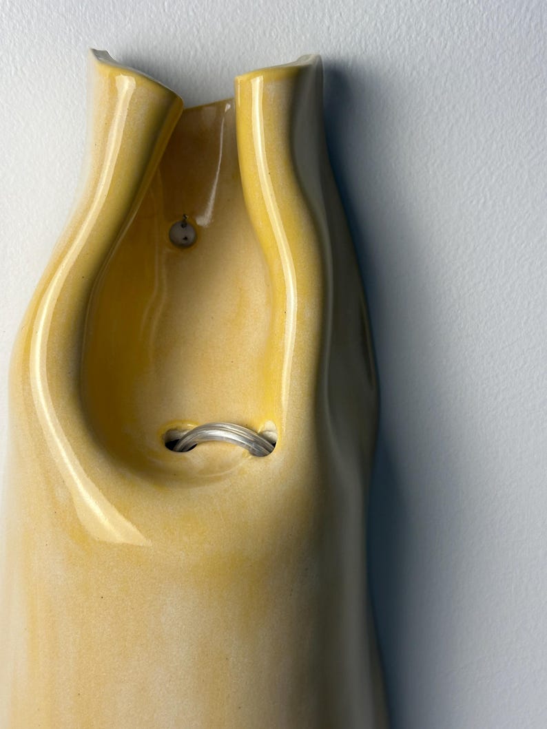 Pu&ograve; includere: Un vaso in ceramica giallo da parete con finitura lucida e forma organica ripiegata. L'oggetto presenta una piccola apertura circolare vicino alla parte superiore e un'apertura centrale pi&ugrave; ampia contenente un anello metallico argentato.