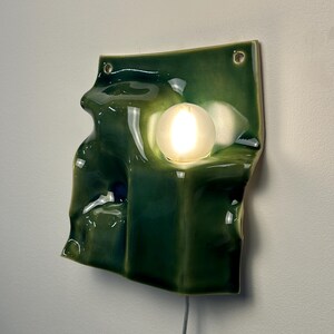 May include: A wall-mounted lamp featuring a glossy, deep green ceramic base with an irregular, undulating surface. The fixture includes a single, spherical, frosted white glass bulb that emits a warm glow, and two small holes at the top for mounting.