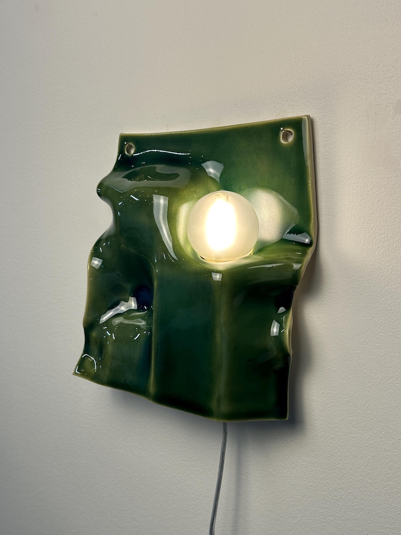 May include: A wall-mounted lamp featuring a glossy, deep green ceramic base with an irregular, undulating surface. The fixture includes a single, spherical, frosted white glass bulb that emits a warm glow, and two small holes at the top for mounting.