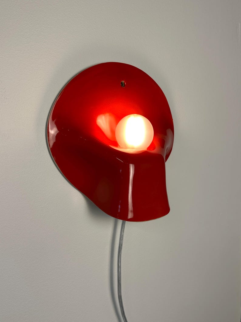May include: A glossy red wall lamp with a curved, organic shape and a single circular opening at the top. A round, frosted white light bulb is positioned in the center, emitting a warm glow. A thin, light-colored power cord hangs from the bottom.
