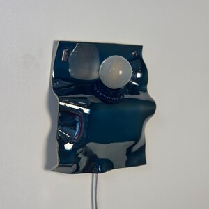 May include: A wall-mounted lamp featuring a reflective, dark blue, wavy metallic plate with an irregular, organic shape. A single, round, frosted white light bulb is mounted centrally on the surface, connected by a thin, light-colored power cord.