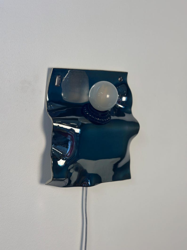 May include: A wall-mounted lamp featuring a reflective, dark blue, wavy metallic plate with an irregular, organic shape. A single, round, frosted white light bulb is mounted centrally on the surface, connected by a thin, light-colored power cord.