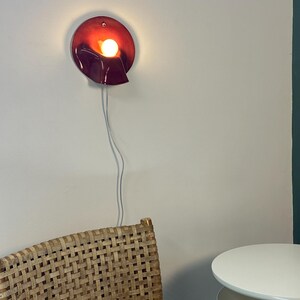 May include: A circular wall lamp with a deep red, glossy finish and a folded, wave-like edge. The lamp features a single glowing light bulb positioned in the centre, emitting a warm light. A thin white power cord hangs vertically from the base.