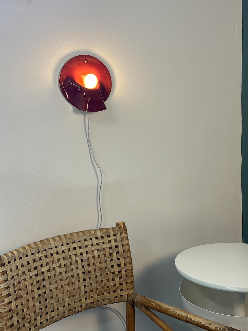 May include: A circular wall lamp with a deep red, glossy finish and a folded, wave-like edge. The lamp features a single glowing light bulb positioned in the centre, emitting a warm light. A thin white power cord hangs vertically from the base.