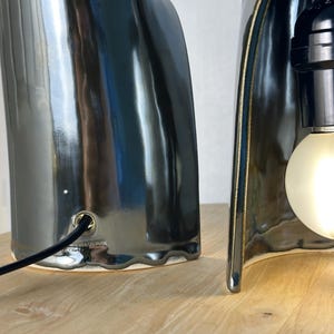 Puede incluir: Dos l&aacute;mparas de mesa esculturales con un acabado de cer&aacute;mica gris carb&oacute;n brillante. Cada l&aacute;mpara presenta una silueta org&aacute;nica curva con un borde de base festoneado. Un cable de alimentaci&oacute;n negro entra por el cuerpo de cer&aacute;mica.