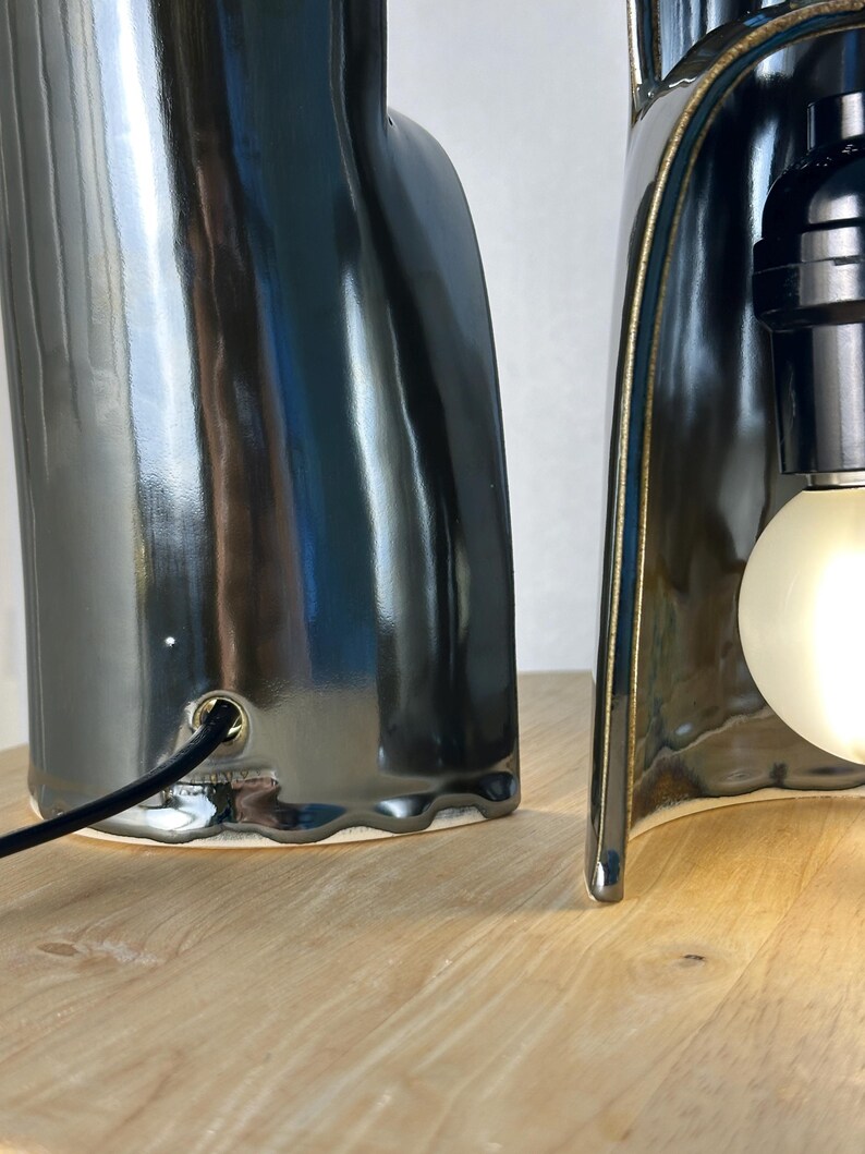 Puede incluir: Dos l&aacute;mparas de mesa esculturales con un acabado de cer&aacute;mica gris carb&oacute;n brillante. Cada l&aacute;mpara presenta una silueta org&aacute;nica curva con un borde de base festoneado. Un cable de alimentaci&oacute;n negro entra por el cuerpo de cer&aacute;mica.