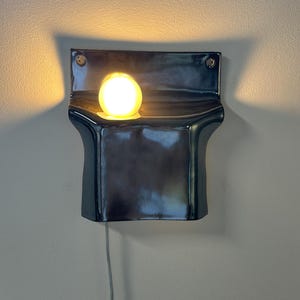 Peut inclure: Une applique murale compos&eacute;e d'un corps en c&eacute;ramique sombre et brillant au design incurv&eacute; en forme de poche. Une ampoule ronde unique, diffusant une lumi&egrave;re chaude, est plac&eacute;e dans l'ouverture sup&eacute;rieure, projetant une lueur douce.