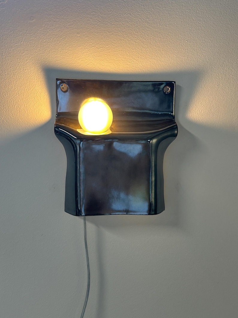Peut inclure: Une applique murale compos&eacute;e d'un corps en c&eacute;ramique sombre et brillant au design incurv&eacute; en forme de poche. Une ampoule ronde unique, diffusant une lumi&egrave;re chaude, est plac&eacute;e dans l'ouverture sup&eacute;rieure, projetant une lueur douce.