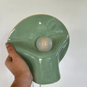 May include: A sage green ceramic wall sconce with a smooth, glossy finish and a wavy, organic shape. The fixture features a central circular opening holding a round, frosted white light bulb and a small hole at the top for mounting purposes.