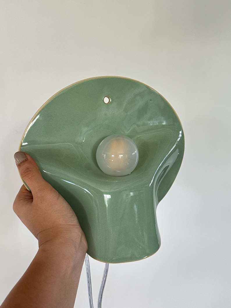 May include: A sage green ceramic wall sconce with a smooth, glossy finish and a wavy, organic shape. The fixture features a central circular opening holding a round, frosted white light bulb and a small hole at the top for mounting purposes.