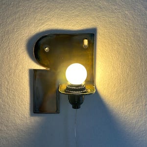 May include: A wall-mounted lamp featuring a glossy, dark-toned ceramic backplate shaped like the letter R. The fixture includes a central socket holding a round, glowing white light bulb, with a thin power cord extending downwards from the base.
