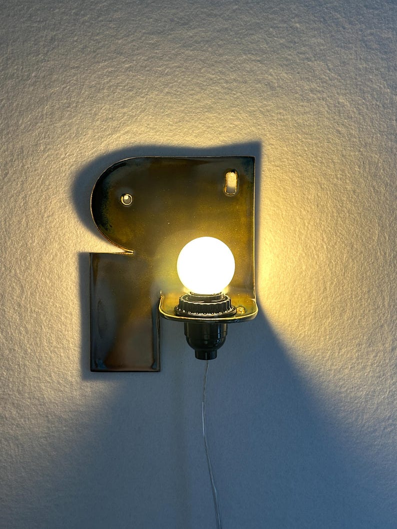 May include: A wall-mounted lamp featuring a glossy, dark-toned ceramic backplate shaped like the letter R. The fixture includes a central socket holding a round, glowing white light bulb, with a thin power cord extending downwards from the base.