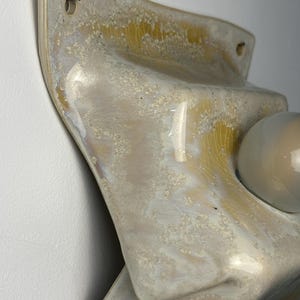 May include: A wall-mounted ceramic light fixture featuring a glossy, textured glaze in shades of cream, beige, and pale yellow. The surface has a crackled, crystalline appearance with a smooth, rounded white glass bulb protruding from the center.